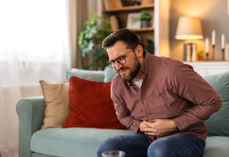 Terrible stomachache. Frustrated handsome young man hugging his belly and keeping eyes closed while lying on the couch at home