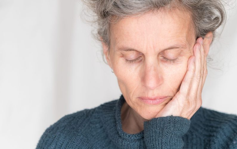 Close up head and shoulders view of older woman in blue sweater with eyes closed and hand on head (selective focus)