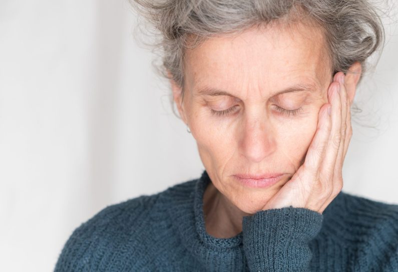 Close up head and shoulders view of older woman in blue sweater with eyes closed and hand on head (selective focus)